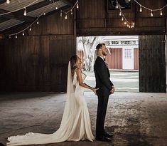 a bride and groom standing in an old barn looking at each other with lights hanging from the ceiling