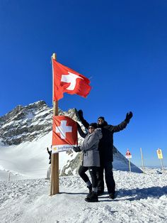 two people standing next to each other in the snow with their arms out and one person holding up a swiss flag