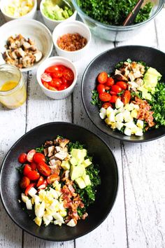 two black plates filled with salad on top of a white wooden table next to bowls of vegetables