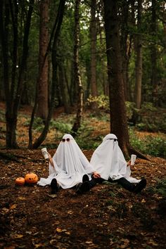 two people dressed in white cloths sitting on the ground with pumpkins and trees