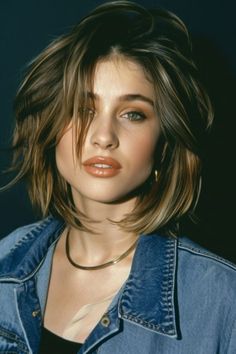 Young woman with tousled brown hair, wearing a denim jacket and gold necklace, against a dark background.