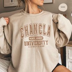 Chancla University sweatshirt is the perfect gift for any Latina mom!  ♣️ Product Details: ▪️ Brand: Experience comfort and style with Gildan 1800. ▪️ Style: Unisex Adult Sizing ▪️ Please Note: Props used in photos are NOT included with purchase. ⚫️ Fabric Material:    ▪️The gildan 18000 is made of 50% cotton & 50% polyester    ▪️(Heather Sport colors: 60% polyester, 40% cotton).     ▪️ Features a comfy unisex fit with a ribbed collar, cuffs, and a waistband with spandex.     ▪️ Made of pill res Casual Slogan Sweatshirt For Mother's Day, Casual Text Print Sweatshirt For Mother's Day, Casual Mother's Day Slogan Sweatshirt, Casual Crew Neck Sweatshirt For Mother's Day, University Crewneck, University Sweatshirts, Favorite Sweater, Brand Experience, Air Jet