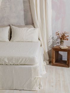 a white bed sitting next to a window with curtains on top of it and a small table in front of it