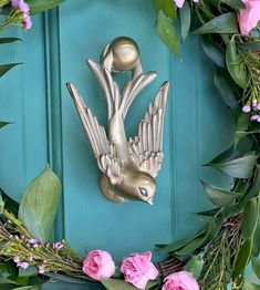 a metal bird on a blue door surrounded by pink flowers and greenery with a gold ball in the center