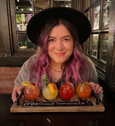 a woman with pink hair is holding a tray full of different types of cocktails