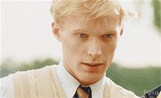 a young man with blonde hair wearing a sweater and tie, looking at the camera