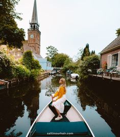 a woman sitting on the bow of a boat in a canal with buildings and trees