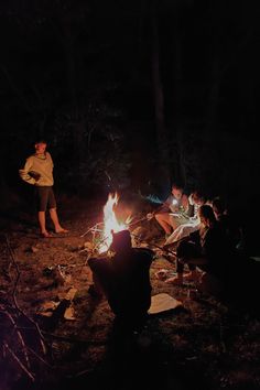 several people sitting around a campfire in the woods at night with one person standing