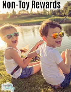 two young children sitting on the grass next to a body of water with fishing rods in their hands