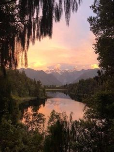 a lake surrounded by trees and mountains under a cloudy sky at sunset in the distance