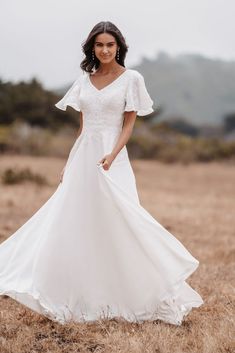 a woman in a white wedding dress standing in a field with her hands on her hips