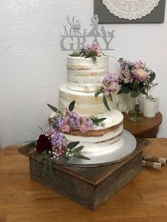 a three tiered cake with flowers and greenery sits on a wooden stand in front of a white wall