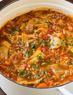 a white pot filled with stew and vegetables on top of a wooden table next to a spoon