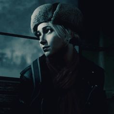 a woman wearing a hat and scarf in front of a window with the city lights behind her