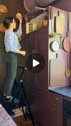 a woman standing on a stepladder in front of a kitchen with pots and pans hanging from the wall