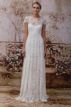 a woman standing in front of a wall wearing a white wedding dress with long sleeves