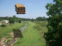 a school bus flying over a river in the middle of a lush green field next to a forest