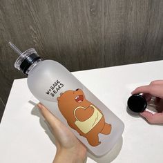 a hand holding a water bottle with a cartoon bear on the front and bottom, next to a person's finger