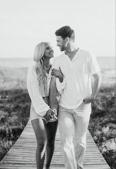 a man and woman walking on a wooden walkway near the ocean in black and white