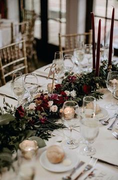 the table is set with white plates and silverware, red flowers, greenery and candles