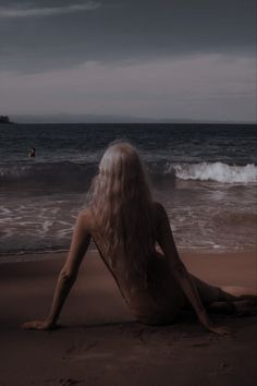 a woman sitting on top of a sandy beach next to the ocean under a cloudy sky