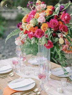 an arrangement of colorful flowers in a tall vase on a table set with place settings