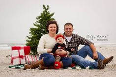 a man and woman are sitting on the beach with their baby in front of a christmas tree