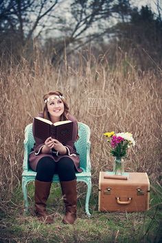 a woman sitting in a chair reading a book next to a suitcase and flower vase