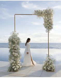 a woman standing in front of an arch with white flowers and greenery on it