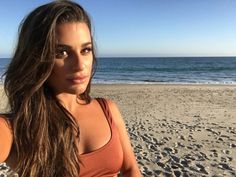 a beautiful young woman standing on top of a sandy beach next to the ocean with her arms behind her back