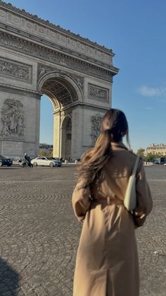 a woman in a trench coat is looking at the arc de trio triumphs on a sunny day