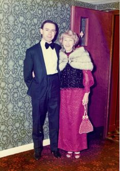 an older couple standing next to each other in front of a wallpapered room