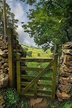 an open wooden gate leading to a stone wall and grassy field with trees in the background