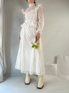 a woman standing in front of a white wall wearing a dress and holding a bouquet