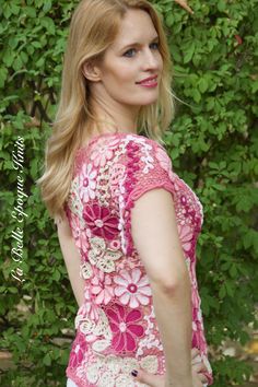 a woman wearing a pink top and white pants standing in front of some bushes with her hand on her hip