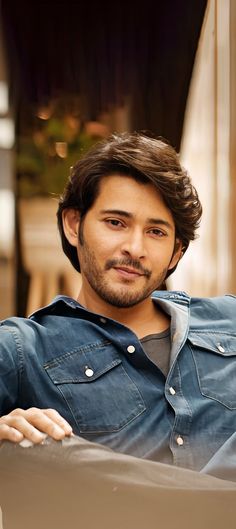 a man with long hair sitting down and looking at the camera while wearing a denim shirt