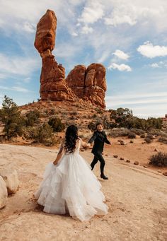 a bride and groom are walking through the desert with rocks in the background at their wedding