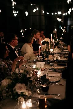 a group of people sitting at a long table with candles in the middle and flowers on it