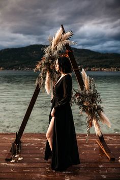 a woman in a black dress standing on a wooden dock next to the ocean with feathers