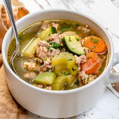 a white bowl filled with soup and vegetables on top of a wooden table next to bread