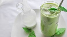 a green drink with ice and mint garnish on a white plate next to a bottle