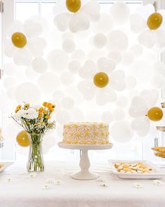 a white table topped with a cake and lots of yellow balloons hanging from the ceiling