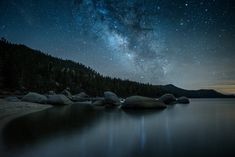 the night sky is filled with stars above some water and rocks in the foreground