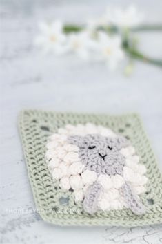 a crocheted square with a white and gray sheep on it next to flowers