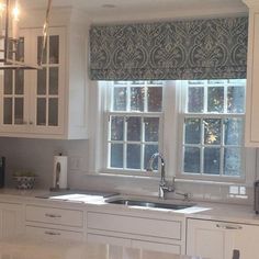 a kitchen with white cabinets and marble counter tops next to a window that has roman shades on it