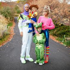 three people in costumes standing on a road with trees and bushes behind them, one holding a child