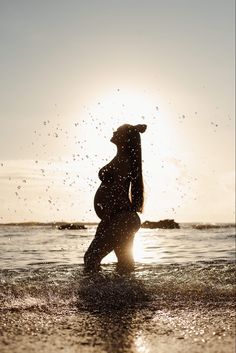 a pregnant woman standing in the ocean at sunset with splashing water all around her