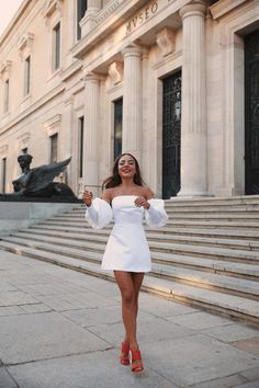 a woman in a white dress is walking down the street with her hand up and smiling