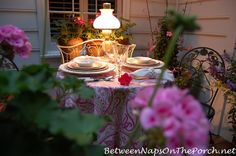 the table is set for two outside with pink flowers and potted plants in the background