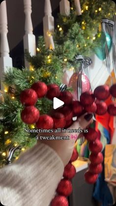 someone is holding up some christmas balls in front of a staircase with garlands and lights on it